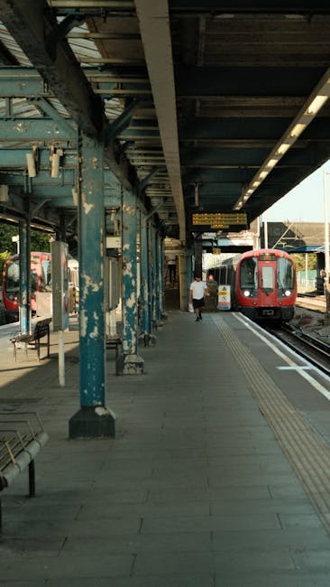 A train station platform with weathered blue-painted metal columns supporting an overhead canopy, which provides shelter for waiting passengers. The platform surface is paved with concrete, featuring 